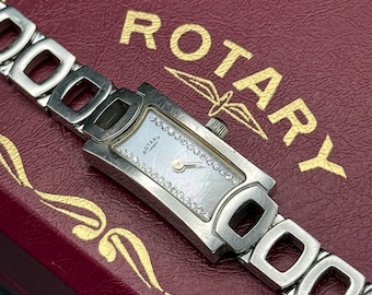 May include: A silver-toned Rotary watch with a rectangular face and a mother-of-pearl dial. The watch is adorned with small, sparkling stones around the face. The watch band has a unique, open-link design. The watch rests on a burgundy box with gold lettering.