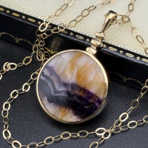 Puede incluir: Un collar dorado con un colgante redondo. El colgante presenta una piedra pulida con tonos morados, amarillos y blancos. La cadena es de estilo eslabón delicado. El collar se muestra sobre una superficie oscura.