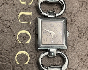 May include: A silver Gucci watch with a square face and a brown face with a GG pattern. The watch has a silver band with a horse bit design.