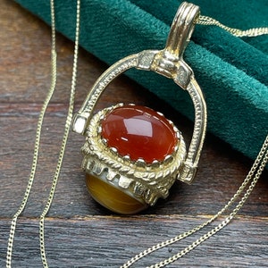 May include: A gold chain necklace with a pendant featuring a red and brown agate stone set in a gold frame. The pendant is designed to rotate.