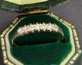 Op de afbeelding: Een gouden ring met zeven kleine, heldere edelstenen in een rij. De ring wordt gepresenteerd in een vintage groene sieradendoos met gouden rand en een groen fluwelen interieur.