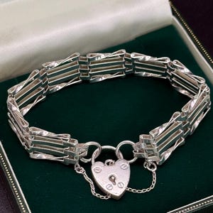 Puede incluir: Una pulsera de plata con un cierre de candado en forma de corazón. La pulsera presenta una serie de eslabones rectangulares conectados por barras retorcidas. El cierre tiene un ojo de cerradura y remaches decorativos, y está unido a la pulsera con una delicada cadena.