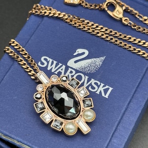 May include: A gold-toned necklace with a large, black, oval-shaped pendant. The pendant is surrounded by clear and gray crystals, and two faux pearls. The necklace is on a blue box with the word "SWAROVSKI" printed on it.