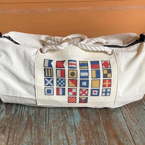May include: A white canvas duffel bag featuring a central rectangular patch printed with a grid of colorful nautical signal flags. The bag includes thick white braided rope handles and a black zipper closure along the top edge of the cylindrical body.
