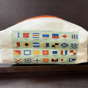 May include: A white zippered pouch featuring a light green rectangular graphic printed with the international maritime signal flags and their corresponding letter names, including Alpha through Zulu, with an orange zipper and grey side tabs.