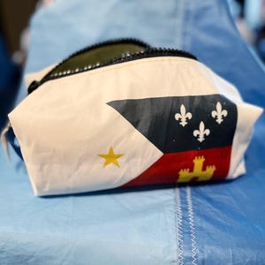May include: A white pouch with a black zipper and a flag design. The flag features a yellow star, three white fleur-de-lis on a black background, and a red and yellow emblem.