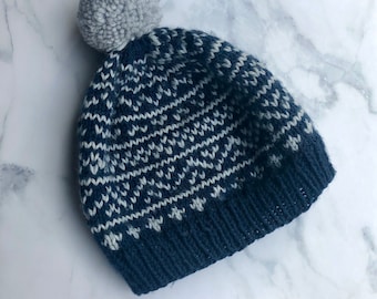 Puede incluir: Un gorro de punto con un patrón geométrico en azul marino oscuro y gris claro. El gorro tiene un borde acanalado en la base y está rematado con un pompón grande y redondo de lana gris claro. El tejido muestra un diseño texturizado repetitivo.