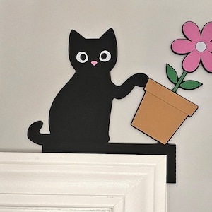 Peut inclure: Une silhouette noire de chat tenant une fleur rose en pot avec des feuilles vertes. Le chat tient le pot avec sa patte droite. Le chat et la fleur sont montés sur un cadre de porte blanc.
