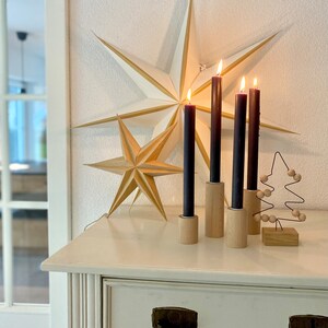May include: Four dark brown candles in wooden holders on a white surface with two gold paper stars in the background.