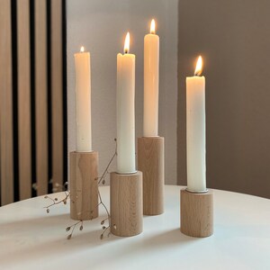 May include: Four white candles burning in wooden candle holders on a white table. The candle holders are made of natural wood and have a simple design.