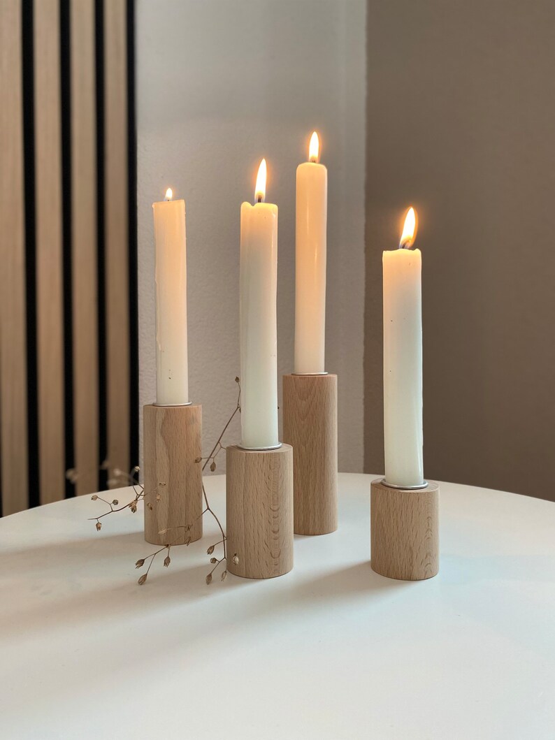 May include: Four white candles burning in wooden candle holders on a white table. The candle holders are made of natural wood and have a simple design.