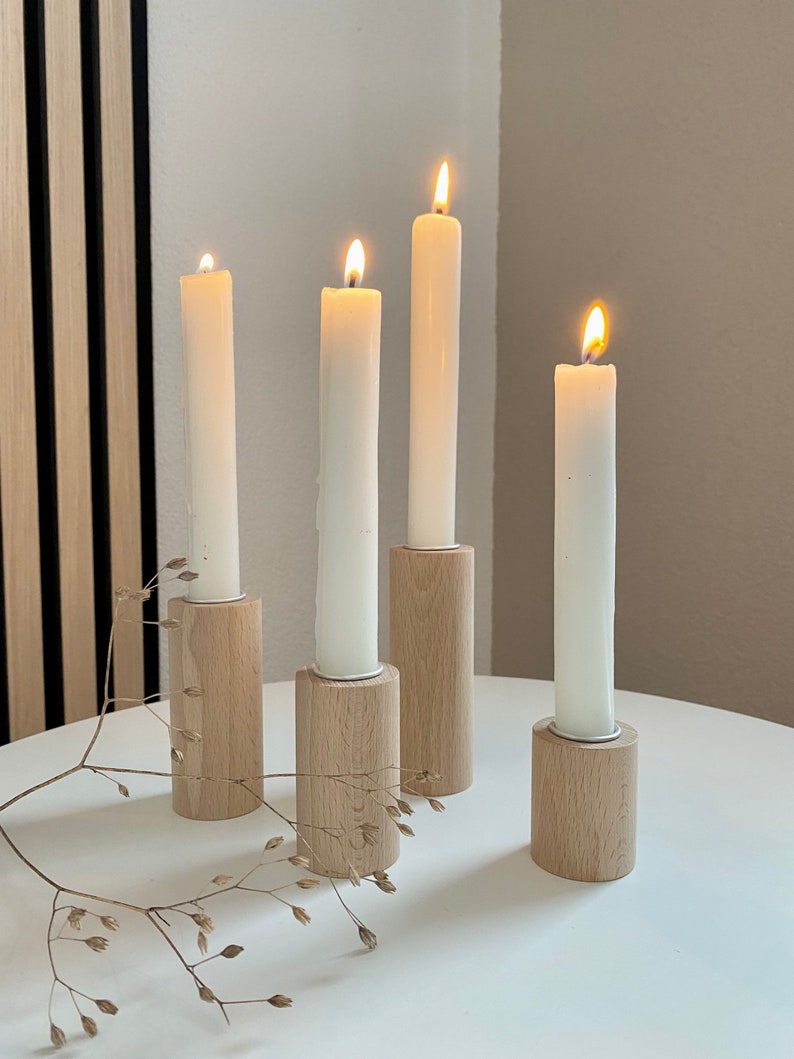 May include: Four white candles burning in wooden candle holders on a white table. The candle holders are made of natural wood and have a simple design.