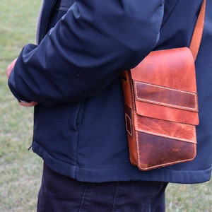 May include: A brown leather crossbody bag with a flap closure and multiple pockets. The bag has a distressed look and is worn over the shoulder of a person.
