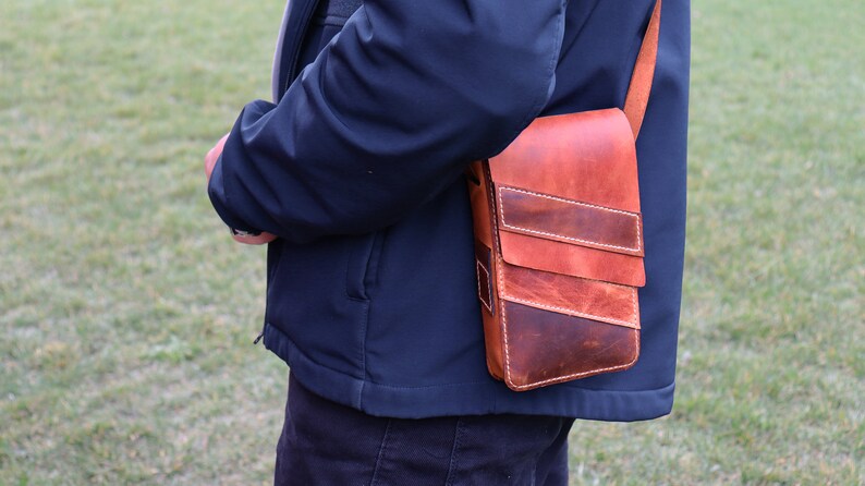 May include: A brown leather crossbody bag with a flap closure and multiple pockets. The bag has a distressed look and is worn over the shoulder of a person.