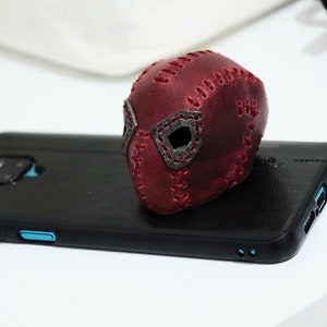 May include: A red leather mask with stitched details and black accents. The mask is sitting on a black smartphone.