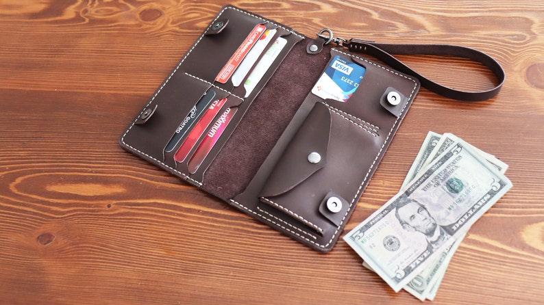 May include: A brown leather wallet with multiple card slots, a large bill compartment, and a smaller pocket. The wallet is open and has several credit cards and a few bills inside. The wallet has a strap for carrying.