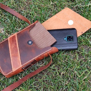 May include: A brown leather phone case with a flap closure and a strap. The case has a woven pattern on the inside and a stitched design on the outside. A black smartphone is inside the case.