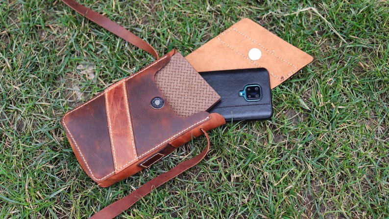 May include: A brown leather phone case with a flap closure and a strap. The case has a woven pattern on the inside and a stitched design on the outside. A black smartphone is inside the case.