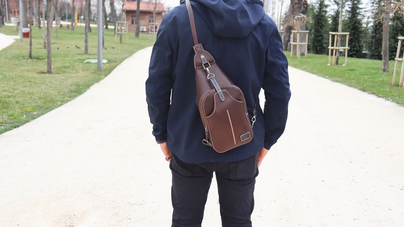 Puede incluir: Un bolso de cuero marr&oacute;n cruzado con una correa que se lleva sobre el hombro de una persona que lleva una chaqueta azul y pantalones vaqueros.