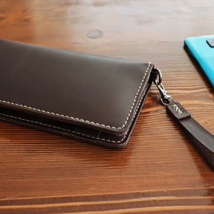 May include: A brown leather wallet with a white stitch detail and a matching brown leather strap. The wallet is lying on a wooden surface next to a blue smartphone.