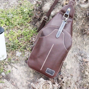Puede incluir: Un bolso de cuero marr&oacute;n cruzado con detalles de costura blanca y una correa negra. El bolso est&aacute; sentado en un tronco de &aacute;rbol con hierba verde y un termo blanco.