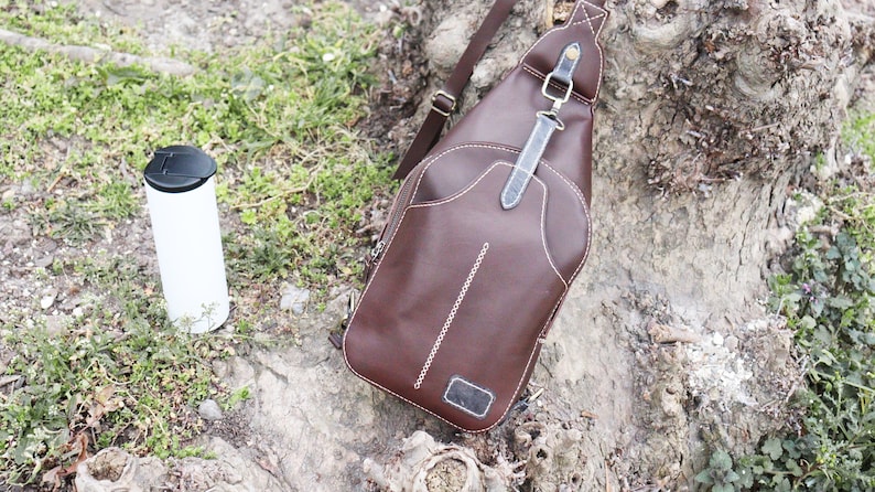 Puede incluir: Un bolso de cuero marr&oacute;n cruzado con detalles de costura blanca y una correa negra. El bolso est&aacute; sentado en un tronco de &aacute;rbol con hierba verde y un termo blanco.