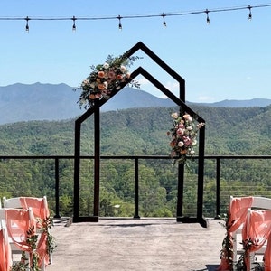 May include: A black metal wedding arch with a geometric design, decorated with pink and peach flowers. The arch is set up on a deck overlooking a mountain range.