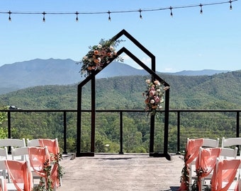 May include: A black metal wedding arch with a geometric design, decorated with pink and peach flowers. The arch is set up on a deck overlooking a mountain range.