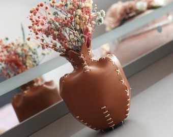 Könnte beinhalten: Eine braune Ledervase mit dekorativen Nähten, gefüllt mit Trockenblumen in Orange-, Rosa- und Gelbtönen. Die Vase steht auf einem Regal, mit einer sichtbaren Spiegelung.