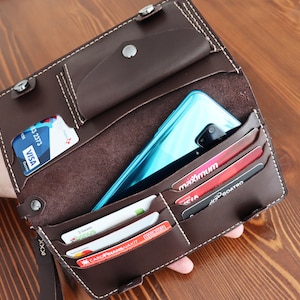 May include: A brown leather wallet with multiple card slots and a phone pocket. The wallet is open and has several cards and a turquoise phone inside.