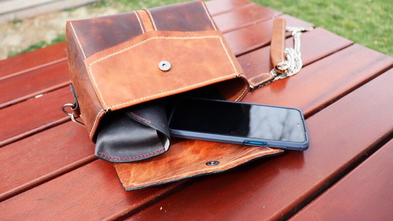 Może przedstawiać: Brązowa sk&oacute;rzana torba na ramię z klapką, otwarta, aby odsłonić czarny smartfon w niebieskim etui i czarne sk&oacute;rzane etui na okulary. Torba leży na drewnianej powierzchni.