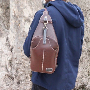 Puede incluir: Un bolso bandolera de cuero marr&oacute;n con cierre de solapa y una correa que se coloca sobre el hombro. El bolso tiene detalles de costura blanca y un peque&ntilde;o bolsillo en la parte delantera.