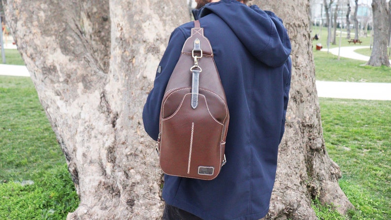 Puede incluir: Un bolso bandolera de cuero marr&oacute;n con cierre de solapa y una correa que se coloca sobre el hombro. El bolso tiene detalles de costura blanca y un peque&ntilde;o bolsillo en la parte delantera.
