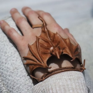 May include: A brown leather bracelet with a sculpted dragon design. The dragon's wings are spread out and the bracelet is tied around a wrist.