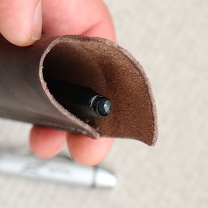 Könnte beinhalten: Ein brauner Leder-Stift-Halter mit einem schwarzen Stift im Inneren. Der Halter ist über den Stift gefaltet, und die Spitze des Stifts ist sichtbar.
