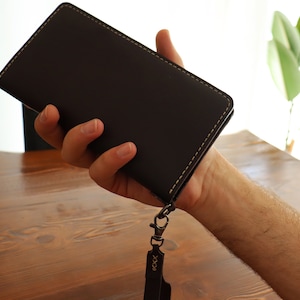 May include: A dark brown leather wallet with a light brown stitch detail and a black metal clasp. The wallet has a black leather strap with the word "XXXX" embossed on it.