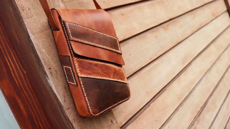 May include: A brown leather crossbody bag with a flap closure and multiple pockets. The bag has a distressed look and is made of high-quality leather.