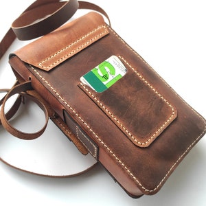 May include: A brown leather crossbody bag with a flap closure and a small pocket on the front. The bag has a long adjustable strap and a card slot on the back.