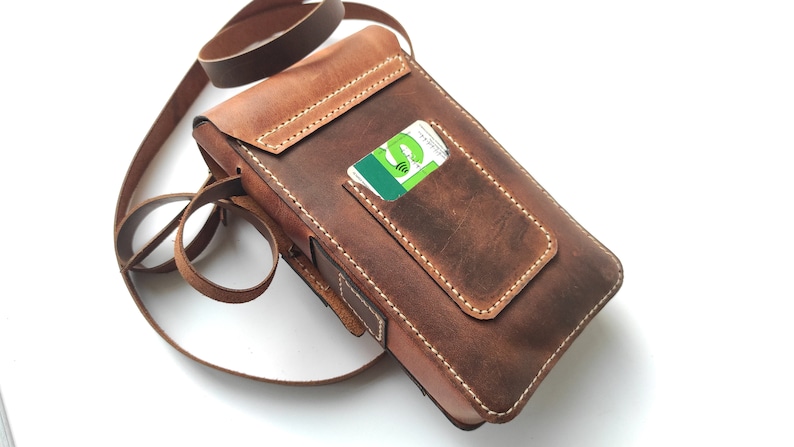 May include: A brown leather crossbody bag with a flap closure and a small pocket on the front. The bag has a long adjustable strap and a card slot on the back.