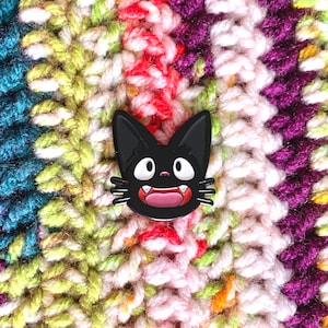 May include: Black cat face enamel pin with white eyes and a pink open mouth. The pin is on a multi-colored crocheted fabric background.