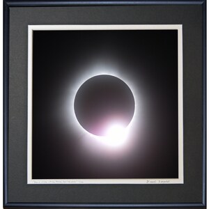 May include: A framed photograph of a total solar eclipse. The sun is completely obscured by the moon, with a bright corona visible around the edges. The image is in black and white, with a slight pink hue to the corona.