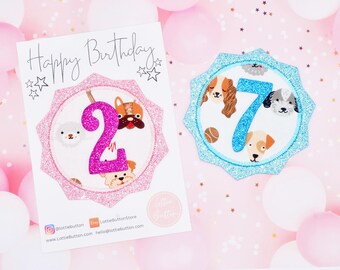 May include: A pink and blue birthday card with a pink glitter number 2 and a blue glitter number 7. The card has a white background with the text "Happy Birthday" and a dog theme.