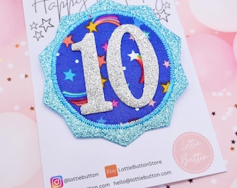May include: A blue and silver glitter fabric patch with the number 10 on it. The patch is shaped like a star and has a blue glitter border. The text "Happy Birthday" is written on a white card behind the patch.