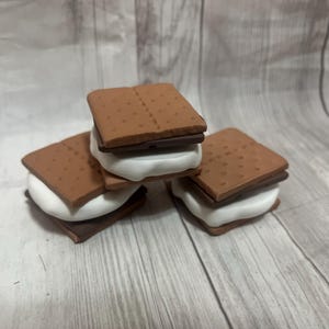 May include: Three miniature s'mores, featuring graham crackers, chocolate, and marshmallow. The graham crackers are a light brown color with dotted details. The chocolate is dark brown, and the marshmallow is white and fluffy.