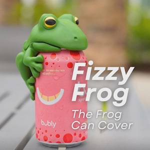 May include: A green frog can cover perched on a pink can of bubbly sparkling water. The can has a smiling face design and red polka dots. Text on the can reads "Fizzy Frog" and "The Frog Can Cover."