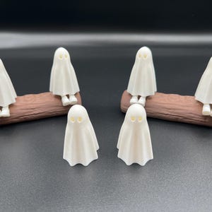 May include: A collection of seven small, white ghost figurines. Six ghosts are perched on two brown, log-shaped bases, while the seventh stands alone. The ghosts have simple, rounded forms with eye-shaped cutouts.