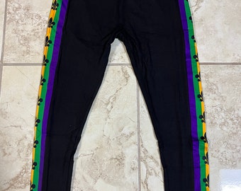 May include: Black leggings with a purple, green and gold stripe down the side. The stripe features a repeating pattern of fleur-de-lis.