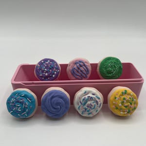 Pode incluir: Um recipiente retangular rosa com oito guloseimas redondas e coloridas. Cada guloseima tem uma base de cor clara coberta com um redemoinho de glacê e granulado em tons de azul, roxo, rosa, verde e amarelo. As guloseimas estão dispostas em duas fileiras.