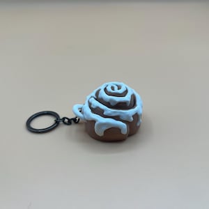 Puede incluir: Un llavero de rollo de canela marrón con glaseado blanco. La miniatura de repostería está unida a un llavero de metal negro. Un accesorio con temática de comida.