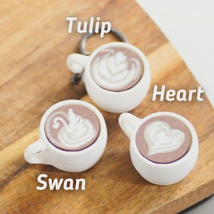 Puede incluir: Tres llaveros de tazas de café blancas con diseños de arte latte: un tulipán, un corazón y un cisne. Cada taza tiene una parte superior marrón color café y un asa blanca. Las palabras "Tulip", "Heart" y "Swan" están escritas en la imagen.